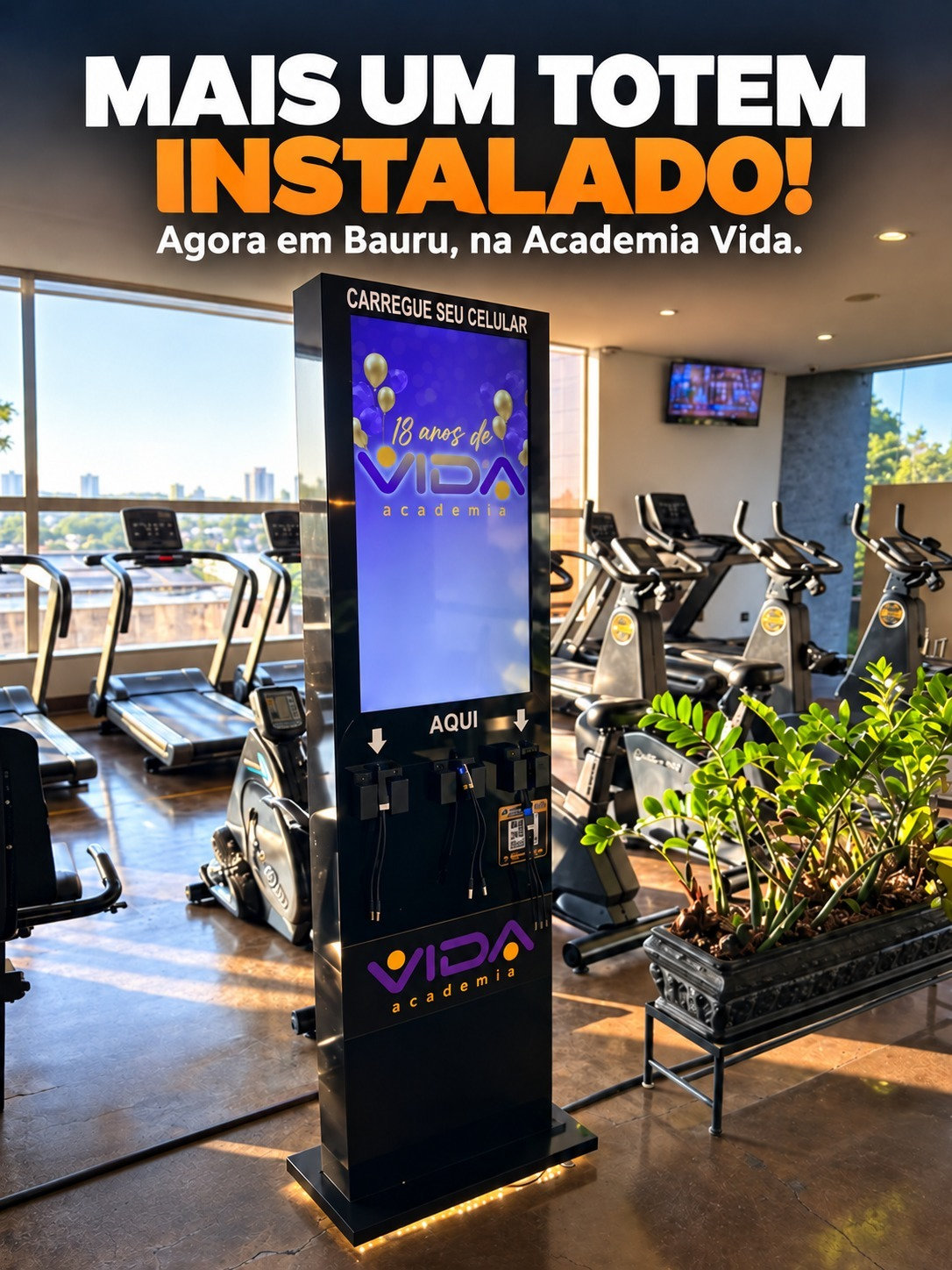 Totem digital com carregadores para celular em espaço comercial, fotografado com duas pessoas ao lado
